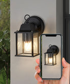 Black exterior wall lamp with clear glass lantern and warm light, shown with smartphone